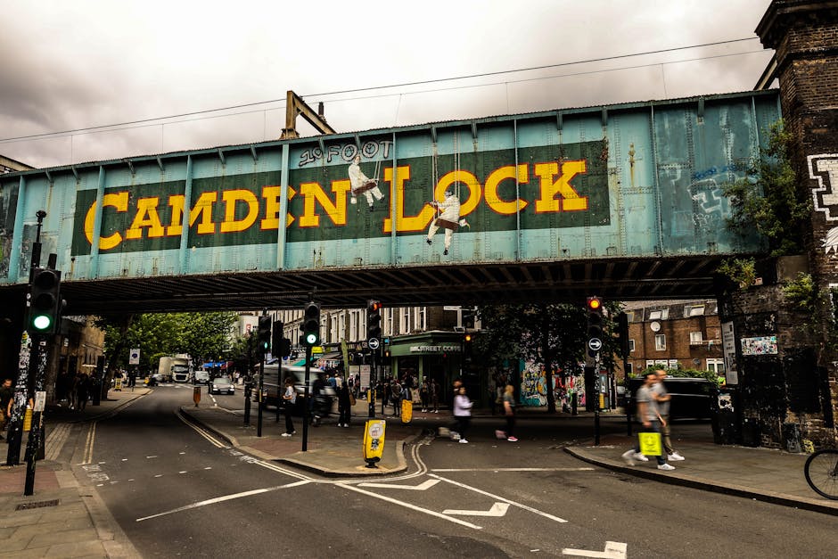 A street scene in Camden displaying a brick building with large arched windows on the left, next to a busy pavement area with pedestrians, some carrying bags or pushing strollers. In the background, a