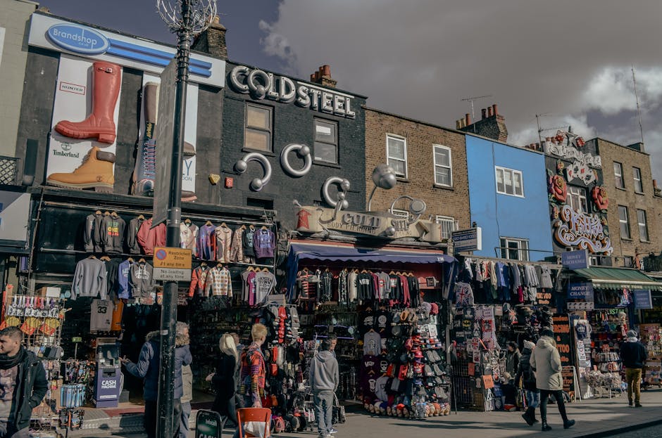 This image shows a busy street scene outside a row of shops, including a clothing store and a signage for brands such as Timberland and Cold Steel, with various merchandise displayed outside on racks 