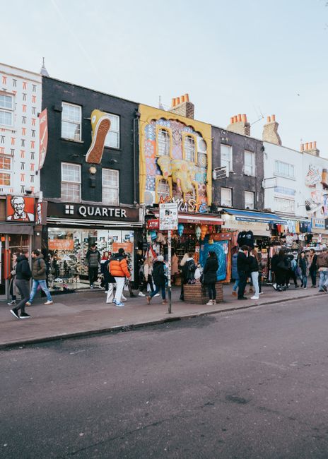 This image shows a busy street scene outside a row of shops, including a clothing store and a signage for brands such as Timberland and Cold Steel, with various merchandise displayed outside on racks 