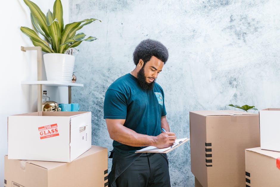 A professional man from Man with Van Camden is indoors during a house relocation, standing amidst several packed cardboard boxes with black tape securing the flaps, some labeled with red 'FRAGILE GLAS