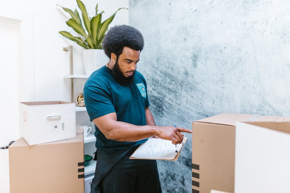 A professional man from Man with Van Camden is indoors during a house relocation, standing amidst several packed cardboard boxes with black tape securing the flaps, some labeled with red 'FRAGILE GLAS